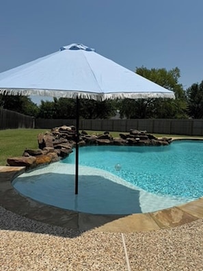 A swimming pool with a stone edge, a rock feature, and a blue umbrella. - Gig'em Getaway Pool House - Sleeps 10 (Bryan)