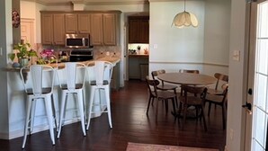 A kitchen with wooden cabinets, a dining area with a round table and chairs, and a hanging light fixture. - Gig'em Getaway Pool House - Sleeps 10 (Bryan)