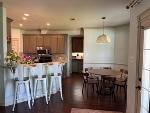 A kitchen with wooden cabinets, a dining area with a round table and chairs, and a hanging light fixture. - Gig'em Getaway Pool House - Sleeps 10 (Bryan)