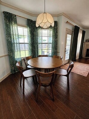 A dining area with a round wooden table and six chairs, a hanging light fixture, and a view of a garden through large windows with green curtains. - Gig'em Getaway Pool House - Sleeps 10 (Bryan)