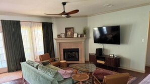 A living room with a sofa, chairs, a coffee table, a fireplace, a television, and a ceiling fan. - Gig'em Getaway Pool House - Sleeps 10 (Bryan)