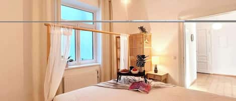 Ein Schlafzimmer mit Bett, Nachttisch, Lampe, einem Fenster mit Vorhängen und einer Tür zu einem anderen Raum.