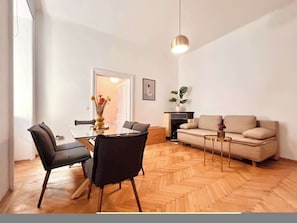 Ein modernes Wohnzimmer mit Essbereich, mit Fischgrätparkett, einer Couch, einem Couchtisch und einer Pendelleuchte.