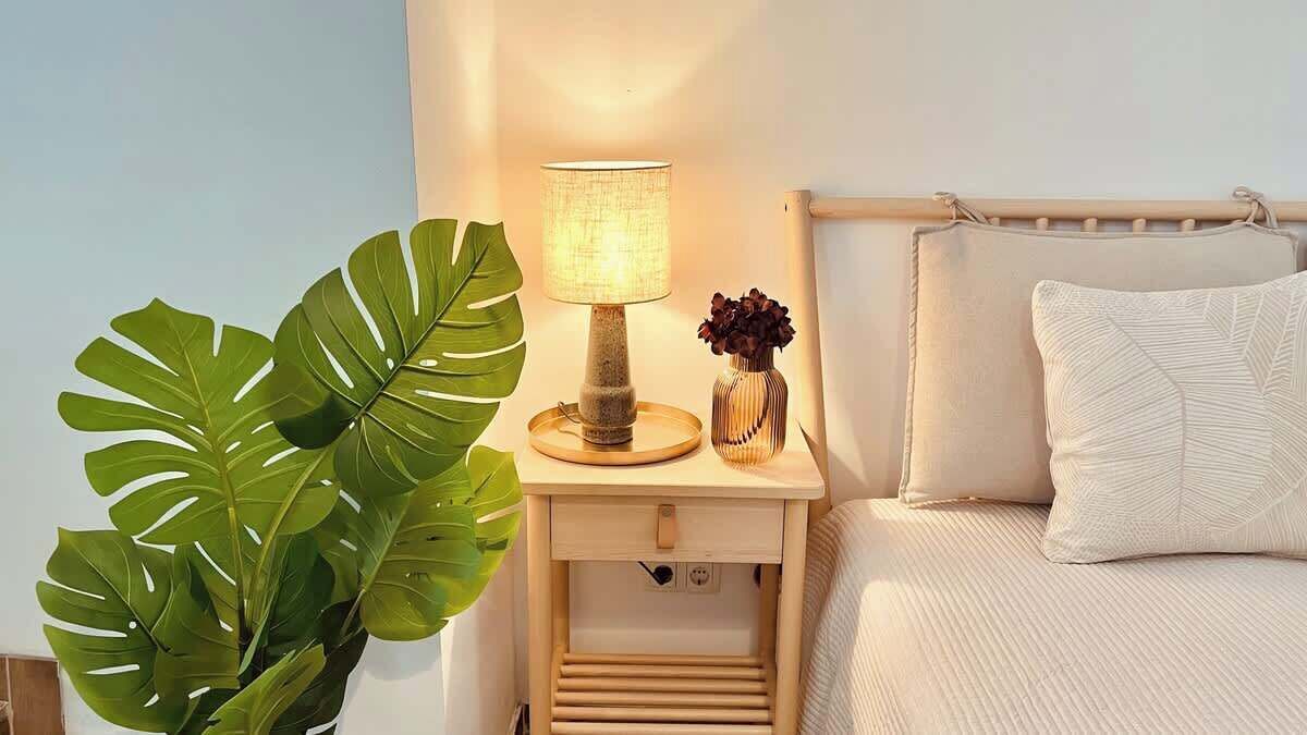 A bedroom with a bedside table, a lamp, a potted plant, and a bed with a pillow.