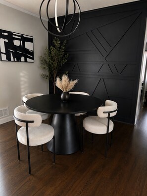 Ein moderner Essbereich mit einem schwarzen Tisch, vier gepolsterten Stühlen, einer Vase mit Trockenpflanzen und einer Wand mit geometrischem Muster.