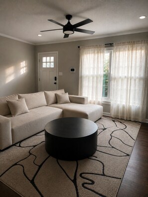 A living room with a sectional sofa, a round coffee table, a ceiling fan, and a window with sheer curtains. - 3BR ModernHilltop Home 5 mins to Atl Airport (Atlanta)