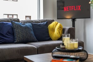 A living room with a grey sofa, blue and yellow cushions, a flat-screen TV displaying 'NETFLIX', and a tray with glasses and a bottle. - Contractor Accommodation in Cannock - Sleeps 5 (Cannock)