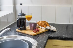 A kitchen counter with a coffee maker, a glass of orange juice, a purple mug, a croissant, and a bunch of grapes. - Contractor Accommodation in Cannock - Sleeps 5 (Cannock)