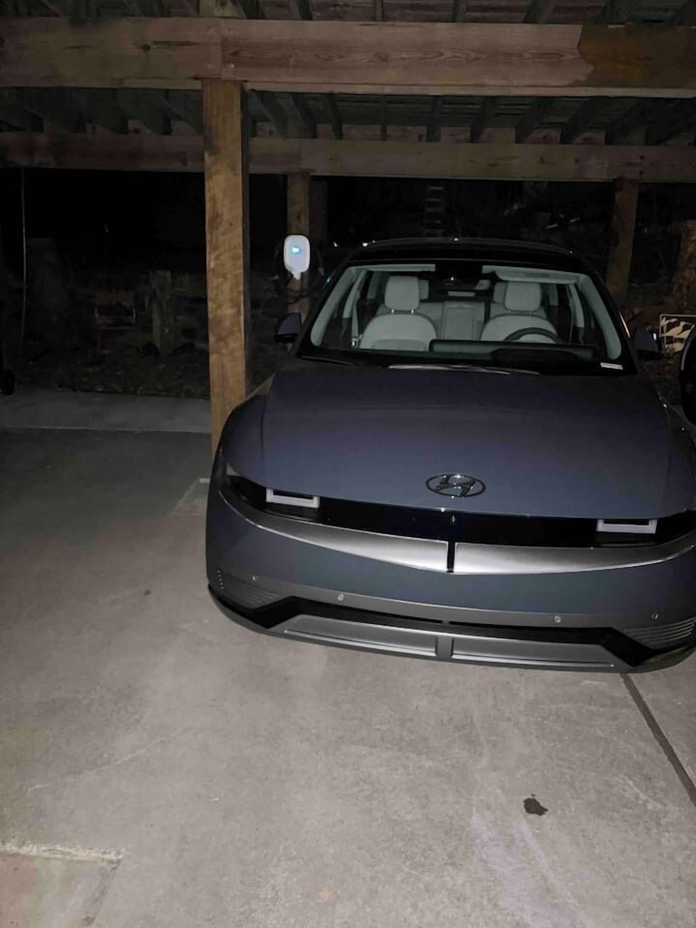Um carro estacionado em uma garagem com pouca luz, piso de concreto e vigas de madeira.