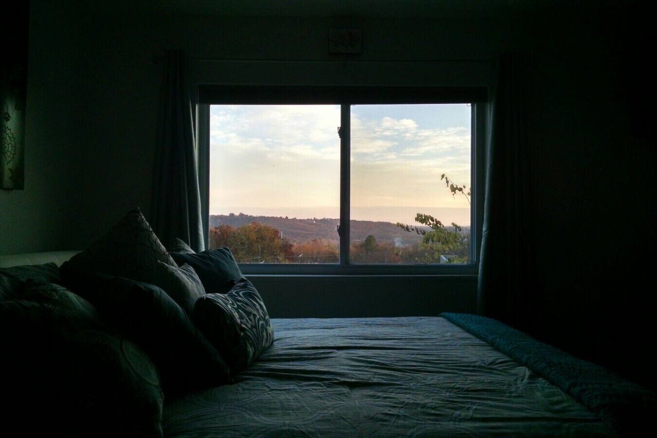 Quarto com uma grande janela, uma cama com almofadas e vista para uma paisagem.