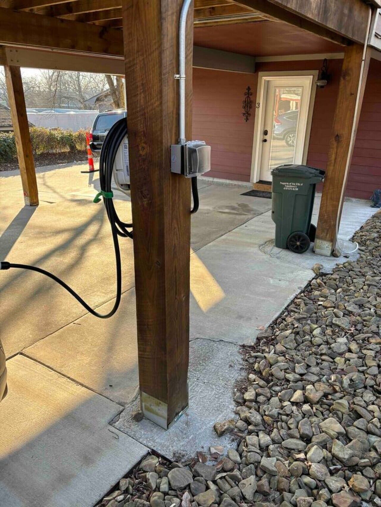 A wooden post with a cable and a meter box, a driveway, a door, and a trash bin.