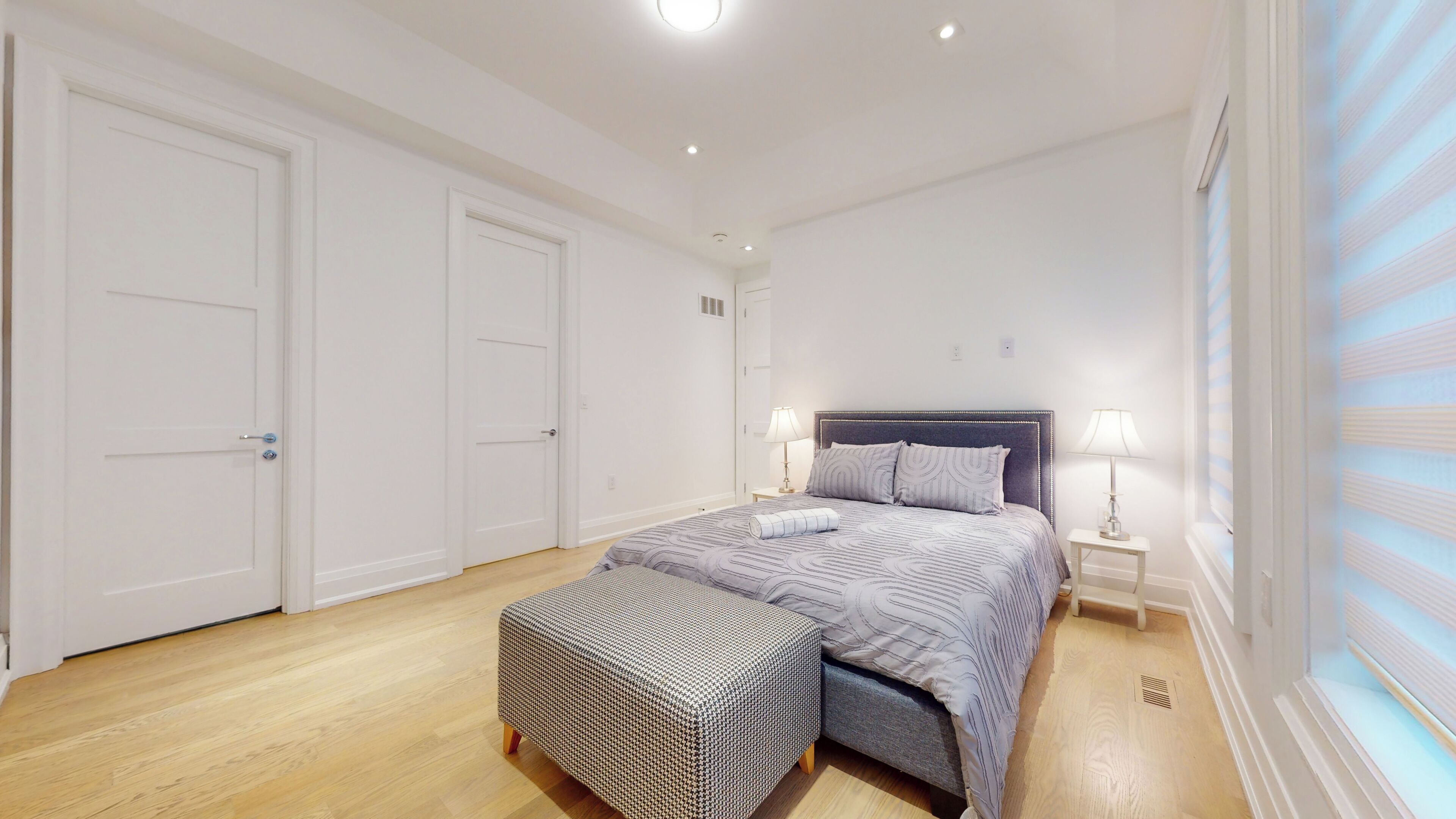 A bedroom with a bed, two bedside tables, a bench, and a large window with blinds.