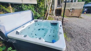 A hot tub with a control panel, surrounded by a gravel area, trees, and a wooden fence.