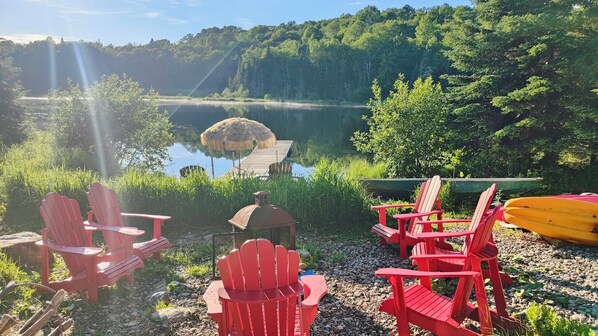 A lakeside setting with red Adirondack chairs, a thatched-roof structure, and a canoe.
