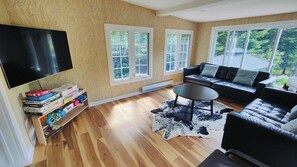 A living room with a flat-screen TV, a bookshelf with board games, a black leather sofa, a round coffee table, and a large window with a view of trees.