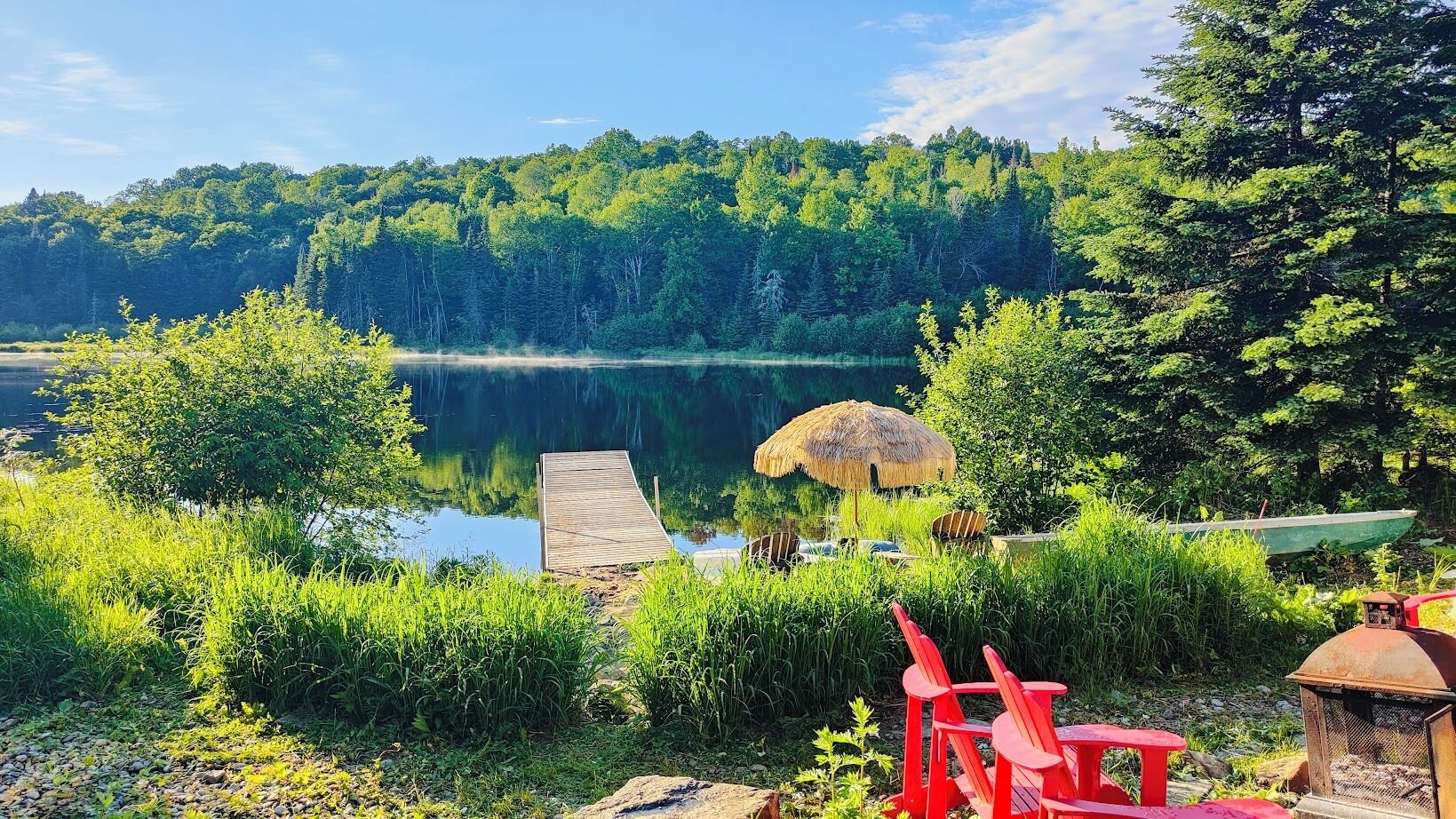 Un cadre idyllique au bord du lac, avec un quai, un parasol en chaume et des chaises rouges.