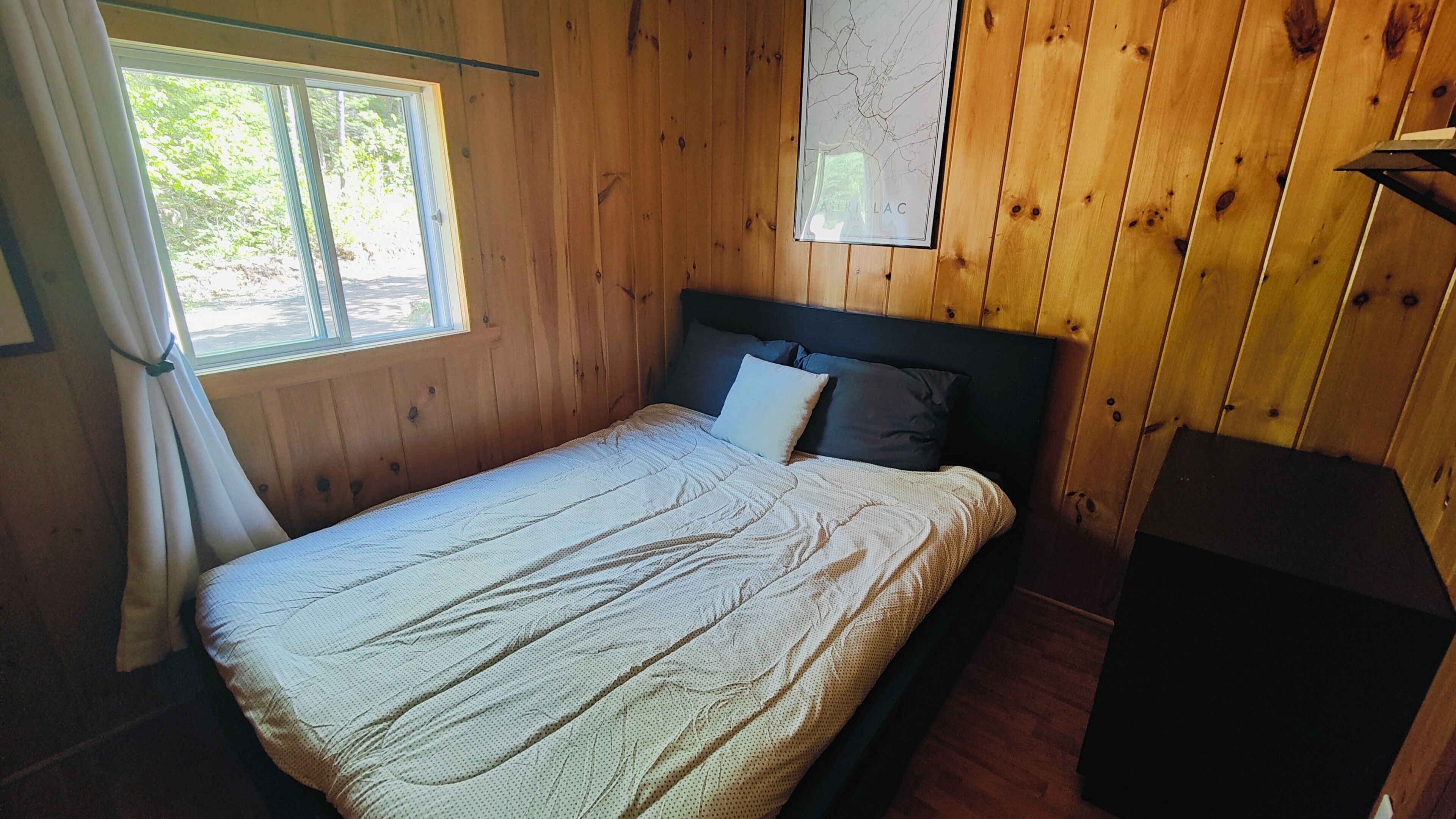 A wooden-paneled room with a bed, a window with curtains, and a map on the wall.