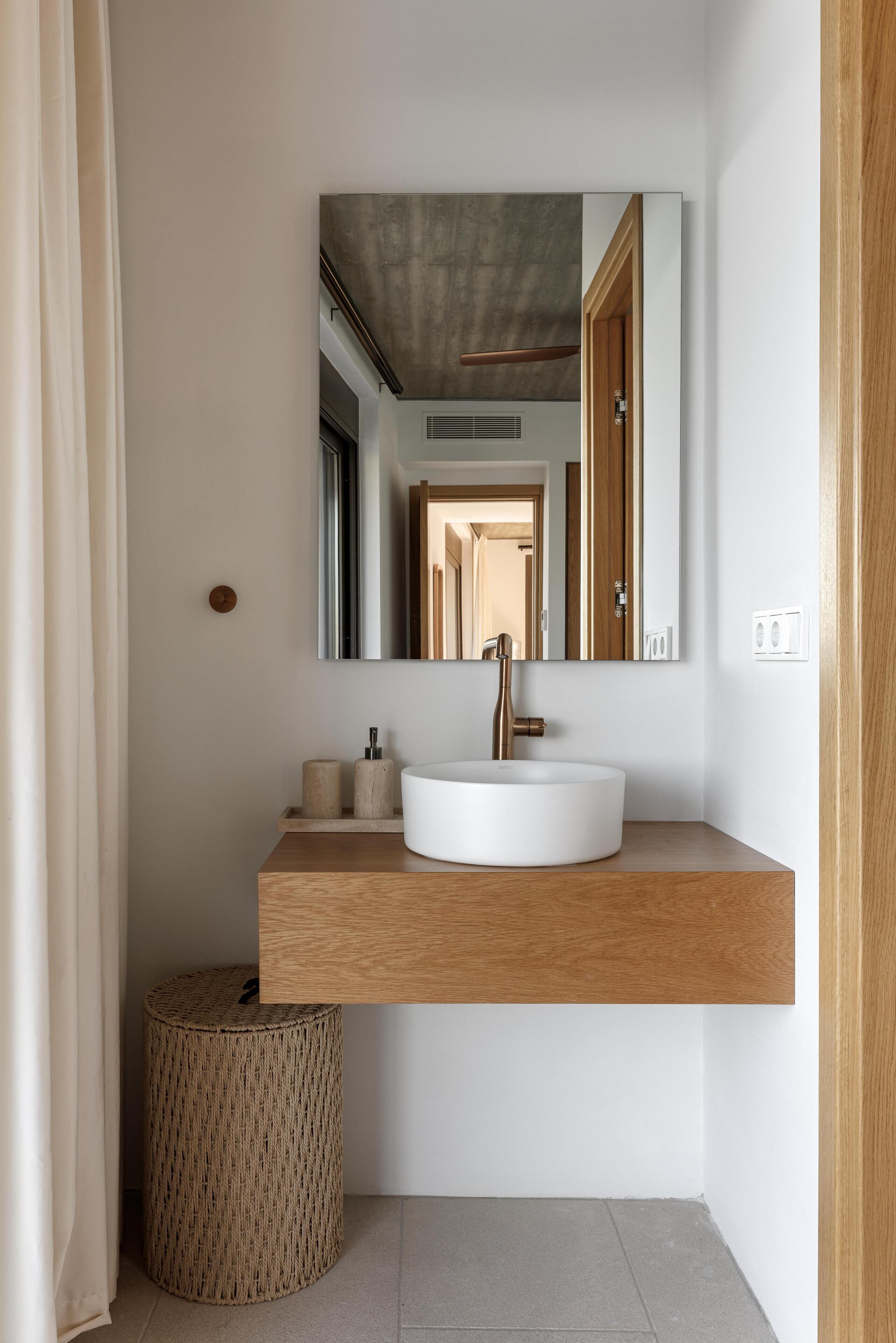 Ein Badezimmer mit einem weißen Waschbecken, einem Holz-Waschtisch, einem runden Korbstuhl, einem Spiegel und einer Tür.