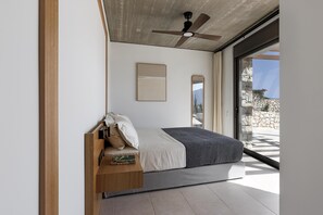 A modern bedroom with a bed, a wooden bedside table, a ceiling fan, and a view through a glass door. - 5 Elements Fiskardo - Two Villas Private Estate (Fiskardo)