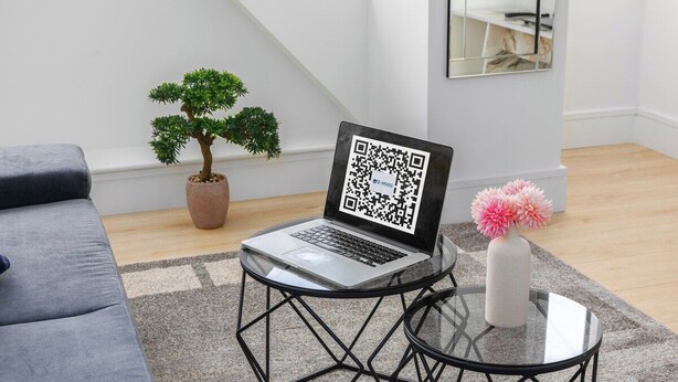 Un portatile su un tavolo di vetro con un codice QR, un bonsai, un vaso con fiori rosa e un divano grigio.