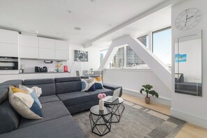 Ein zeitgemäßer Wohnraum mit einem grauen Ecksofa, einem runden Couchtisch, einer Topfpflanze und einem großen Fenster mit Blick auf die Stadt.