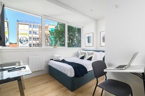 Ein Schlafzimmer mit Bett, Schreibtisch, Stuhl, einem Laptop und einem Fenster mit Blick auf Gebäude und Bäume.