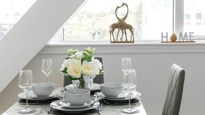 Una tavola imbandita con piatti bianchi, ciotole e bicchieri da vino, un vaso con fiori bianchi e una decorazione a forma di giraffa su un davanzale.