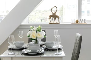 Ein Esstisch ist gedeckt mit weißen Tellern, Schüsseln und Weingläsern, eine Vase mit weißen Blumen sowie eine Giraffenfigur auf einem Fensterbrett.