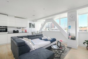 Ein modernes Wohnzimmer mit Schlafsofa, kleinem Tisch mit Laptop und Blick auf die Stadt durch große Fenster.