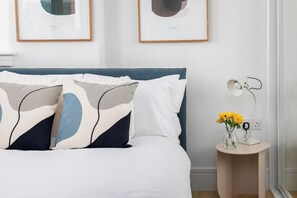 Ein ordentlich gemachtes Bett mit blauem Kopfteil, ein Nachttisch mit Lampe und eine Vase mit Blumen.