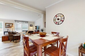 A dining area with a wooden table and chairs, a wall clock, and a living room in the background. - Blackhawk Retreat- Near Lakes, Trails & Golf (Arkdale)