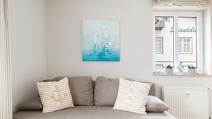 A neutral-toned sofa with striped pillows, a wall-mounted painting of sailboats, and a window with a view of a building and a balcony. - Apartment 'Haus Buskam 29 - Räder Erlebnisbad' with Sea View, Balcony and Wi-Fi (Göhren)