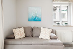Ein sofas in neutraler Farbe mit gestreiften Kissen, ein an der Wand befestigtes Gemälde mit Segelbooten und ein Fenster mit Blick auf ein Gebäude und einen Balkon.