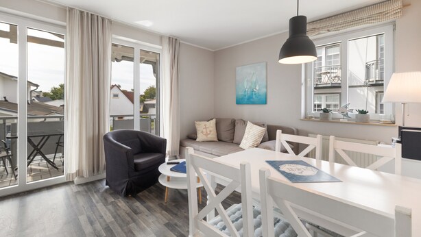 A modern living room with a sofa, a dining table, and a black chair. There is a large window with curtains, a painting on the wall, and a hanging light fixture. - Apartment 'Haus Buskam 29 - Räder Erlebnisbad' with Sea View, Balcony and Wi-Fi (Göhren)