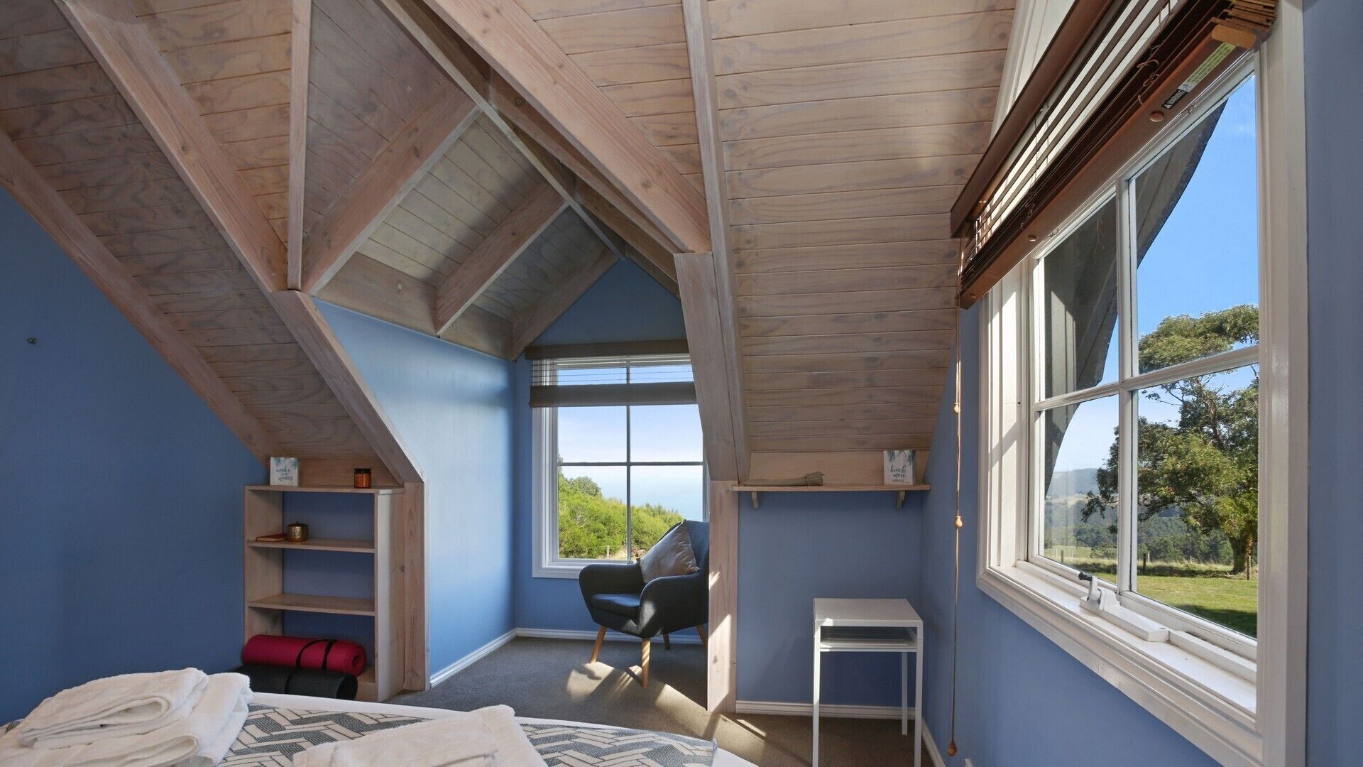 A room with a sloped ceiling, large windows, a bed, a chair, and a small table.