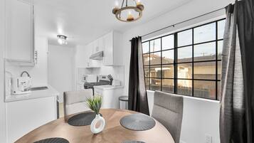 Una cocina moderna con armarios blancos, una mesa de comedor con posavasos negros y vistas a través de un ventanal con cortinas negras.
