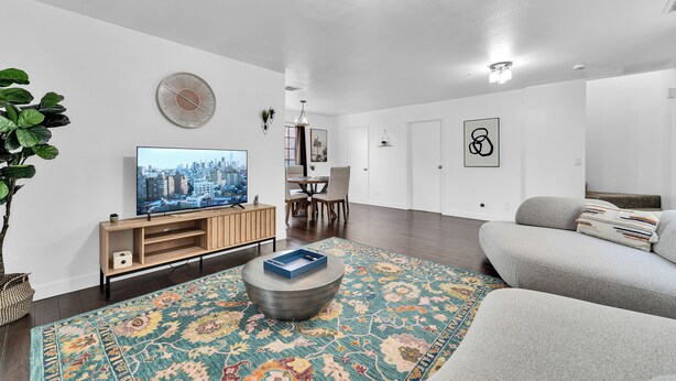 Ein modernes Wohnzimmer mit Flachbildfernseher, rundem Couchtisch, gemustertem Teppich, grauem Sofa und Essbereich mit Stühlen und Tisch.