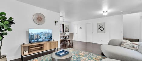 Ein modernes Wohnzimmer mit Flachbildfernseher, rundem Couchtisch, gemustertem Teppich, grauem Sofa und Essbereich mit Stühlen und Tisch.