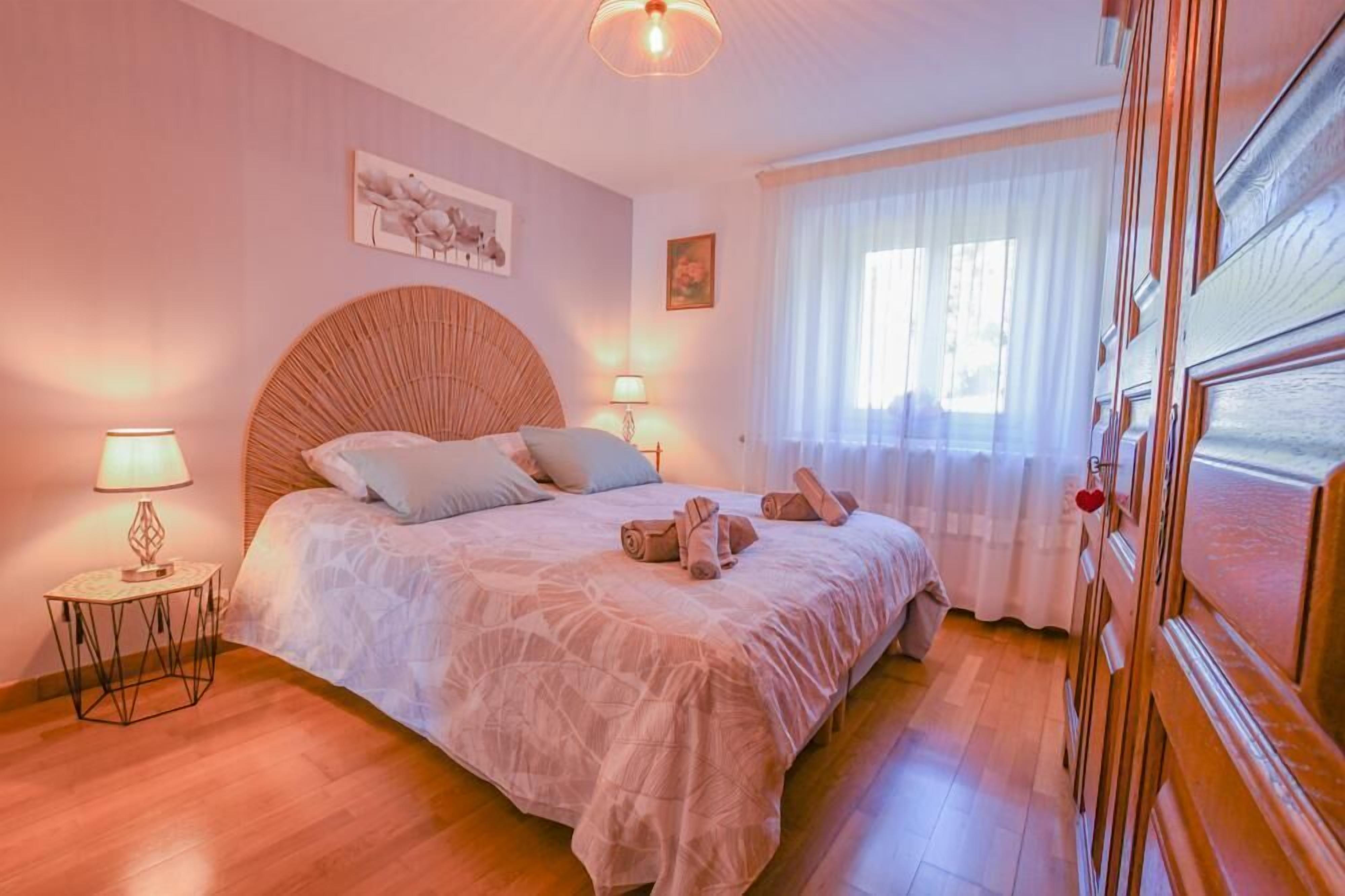 A bedroom with a large bed, two bedside lamps, a round headboard, and a window with curtains.
