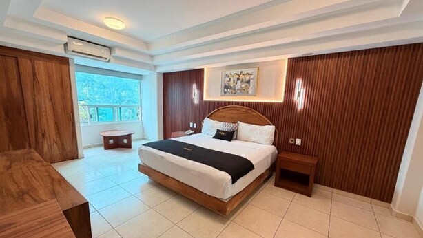 Habitación de hotel con una cama grande, cabecera de madera, una mesita de noche pequeña y una ventana con cortinas.