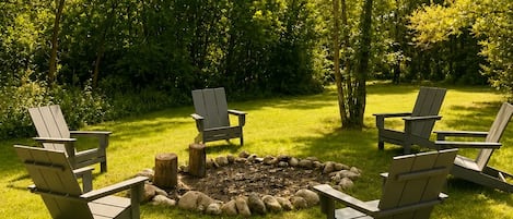 Un hogar con fuego rodeado de sillas Adirondack en un espacio exterior con césped y árboles.
