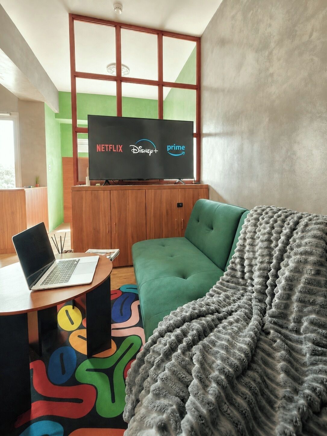 Un salon moderne avec un canapé vert, un plaid gris, un ordinateur portable posé sur une table en bois et un téléviseur affichant les logos de différents services de diffusion en continu.