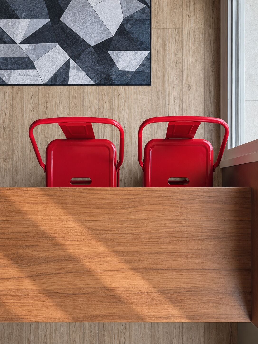 Un mur en bois orné d’un tableau abstrait aux motifs géométriques, ainsi que deux chaises en métal rouge dotées de poignées.