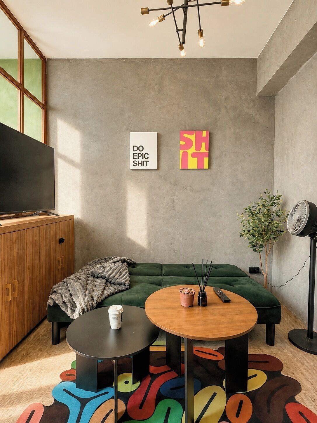 Ein modernes Wohnzimmer mit einem grünen Sofa, einem runden Couchtisch aus Holz, einer schwarzen TV-Konsole und einem Wandventilator.