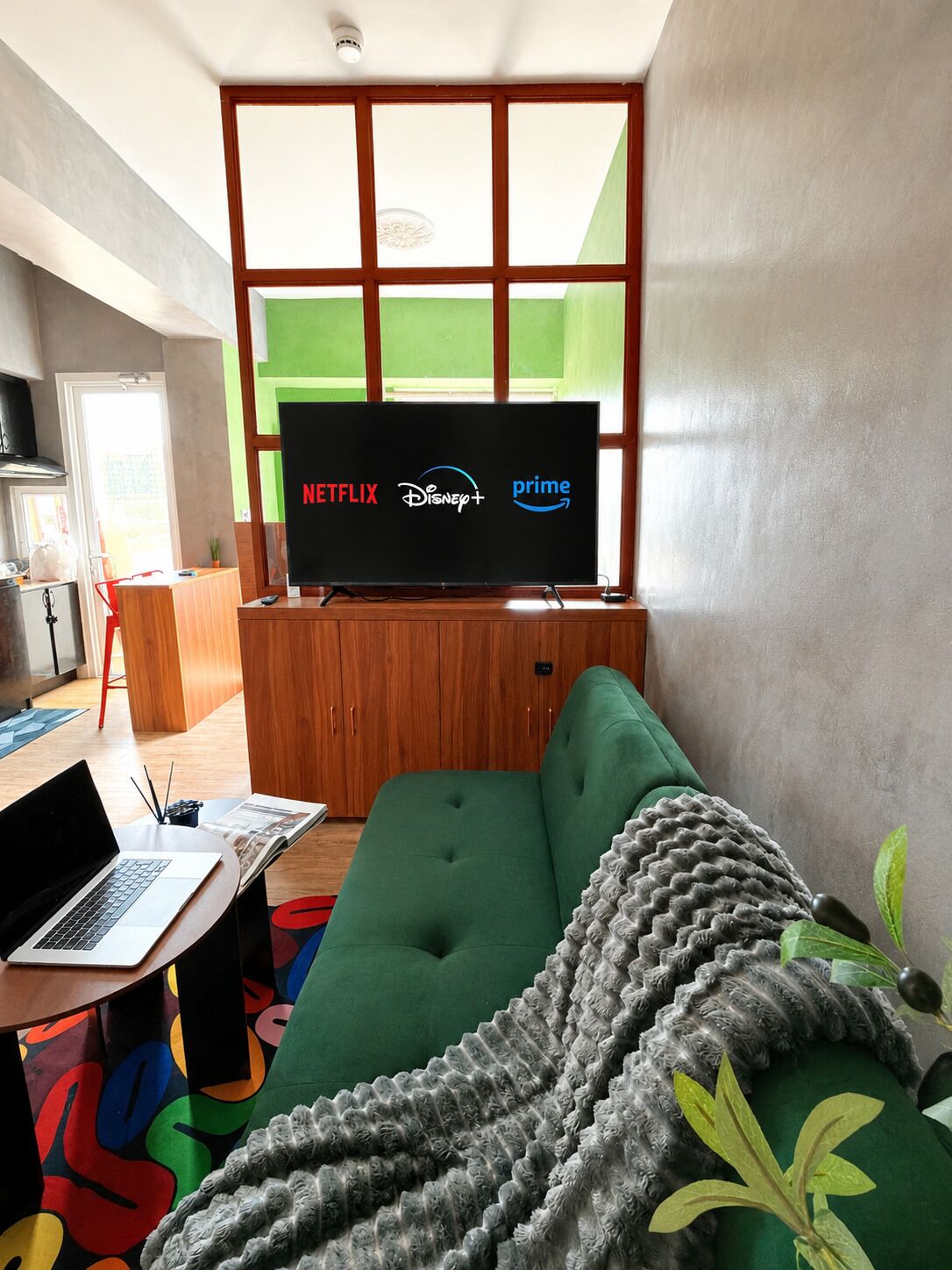 Ein modernes Wohnzimmer mit einem Flachbildfernseher, der Streamingdienste zeigt, einem grünen Sofa, einem Laptop auf einem kleinen Tisch und einem farbenfrohen Teppich.