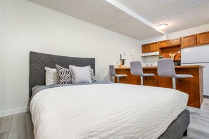 A bedroom with a bed, two chairs, a kitchenette, and a grey headboard.