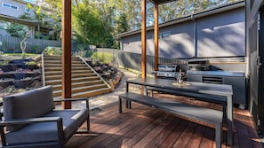 An outdoor deck with a grill, seating, and a staircase leading to a garden area. - Valley Retreat | Luxury Holiday Home (Smiths Lake)