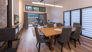 A modern dining area with a wooden table and chairs, a fireplace, and large windows with blinds. - Valley Retreat | Luxury Holiday Home (Smiths Lake)