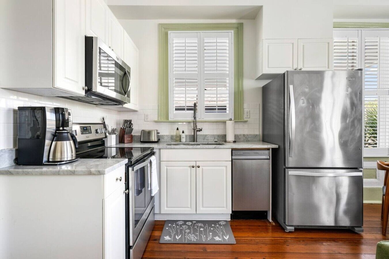 A compact kitchen with white cabinets, a stainless steel refrigerator, a coffee maker, and a window with shutters.