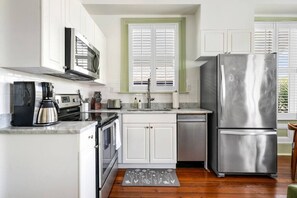 A compact kitchen with white cabinets, a stainless steel refrigerator, a coffee maker, and a window with shutters. - 2BR Spring Street Retreat - B (Charleston)